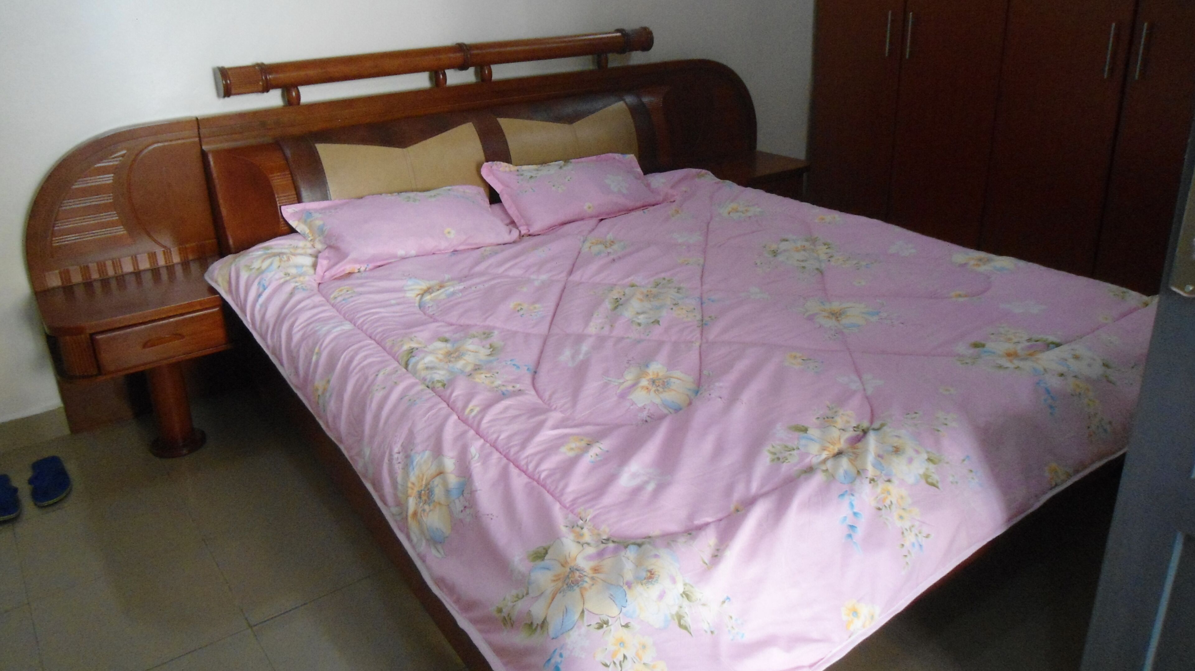 A bedroom with a wooden bed frame, a pink floral bedspread, and a wooden nightstand with a drawer.