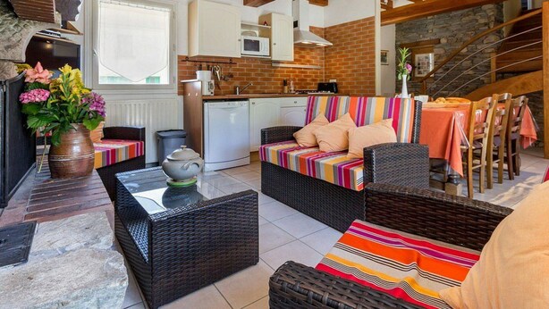 A cozy living room with a sofa, coffee table, and wicker chairs. A kitchen with a microwave and stove is visible in the background.