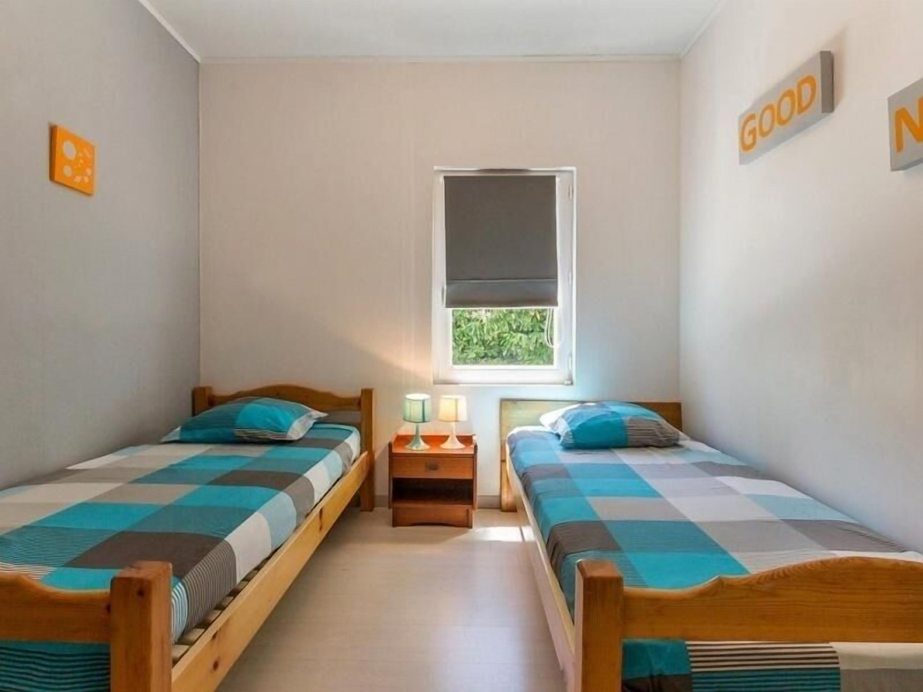 A small, clean room with two single beds, a window, and a wall-mounted sign that reads "GOOD".