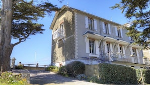 A stone building with a balcony, a clear sky, and a tree in the foreground. - Waterfront 2-Bedroom Cottage with Terrace in Saint-Michel-Chef-Chef (Saint-Michel-Chef-Chef)