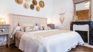 A bedroom with a wooden headboard, a large bed with white bedding, multiple hats on the wall, a fireplace, and a mirror above it.