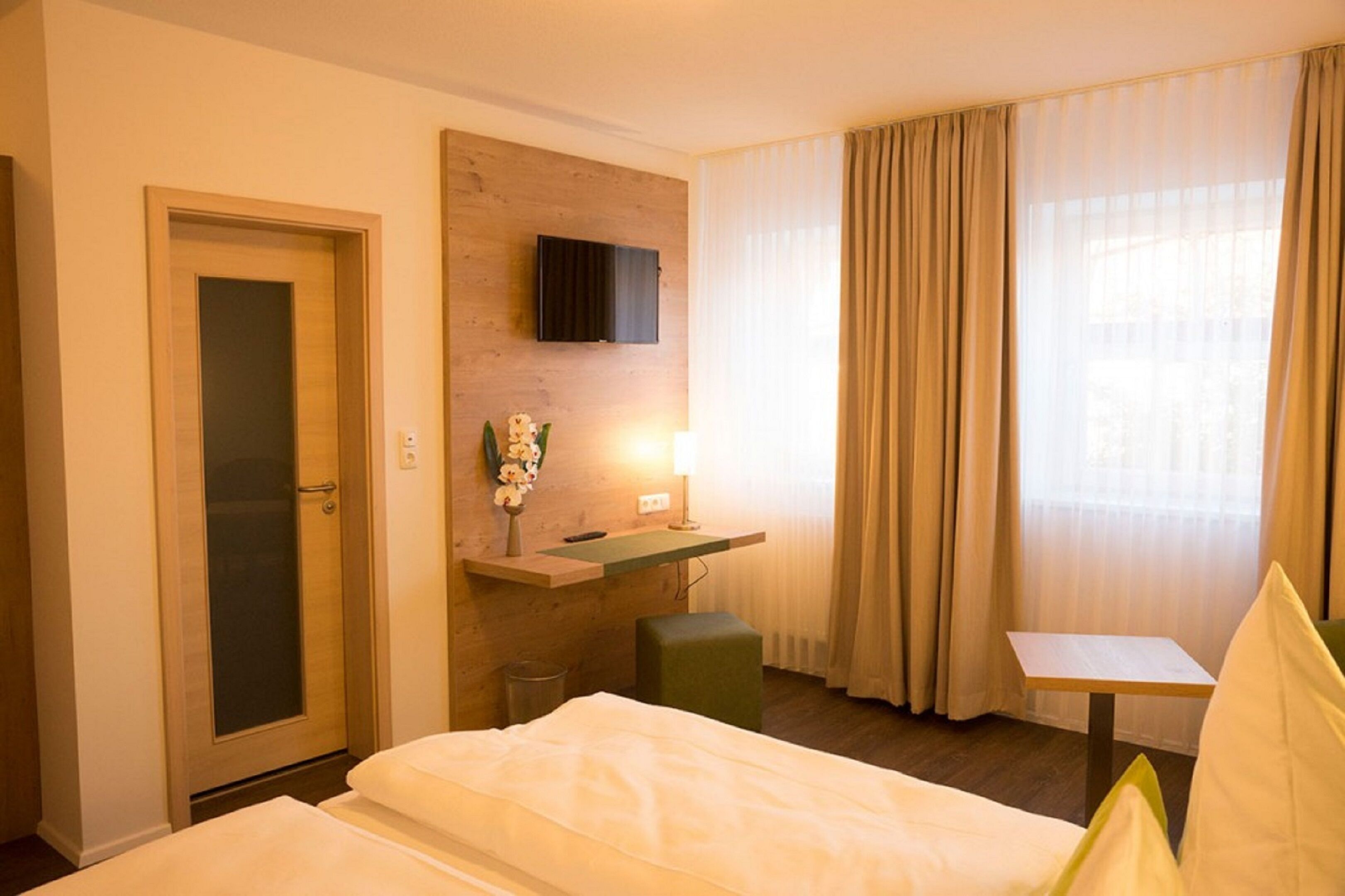 Una habitación de hotel con cama, un escritorio pequeño, televisor montado en la pared, ventana con cortinas y una puerta.