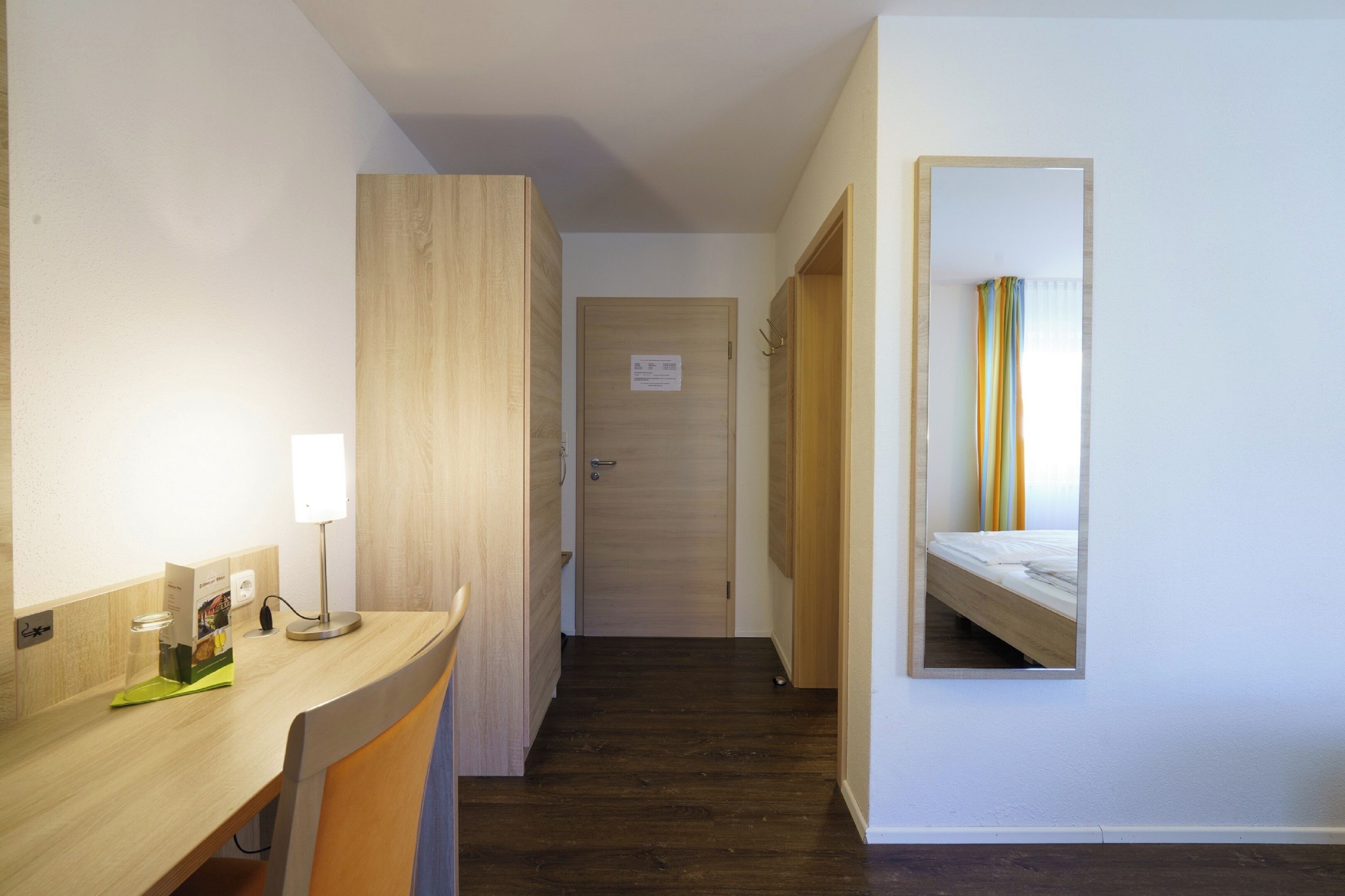 A hotel room with a wooden desk, a mirror, a bed, and a door.
