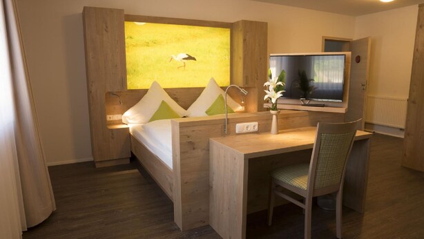 A hotel room with a wooden desk, a bed with a headboard, a television, and a window with a painting of a bird. - Gasthaus Schwarzer Adler (Colmberg)