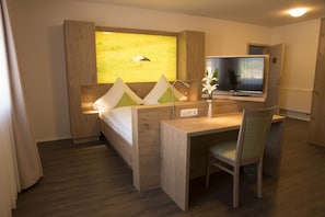 A hotel room with a wooden desk, a bed with a headboard, a television, and a window with a painting of a bird. - Gasthaus Schwarzer Adler (Colmberg)