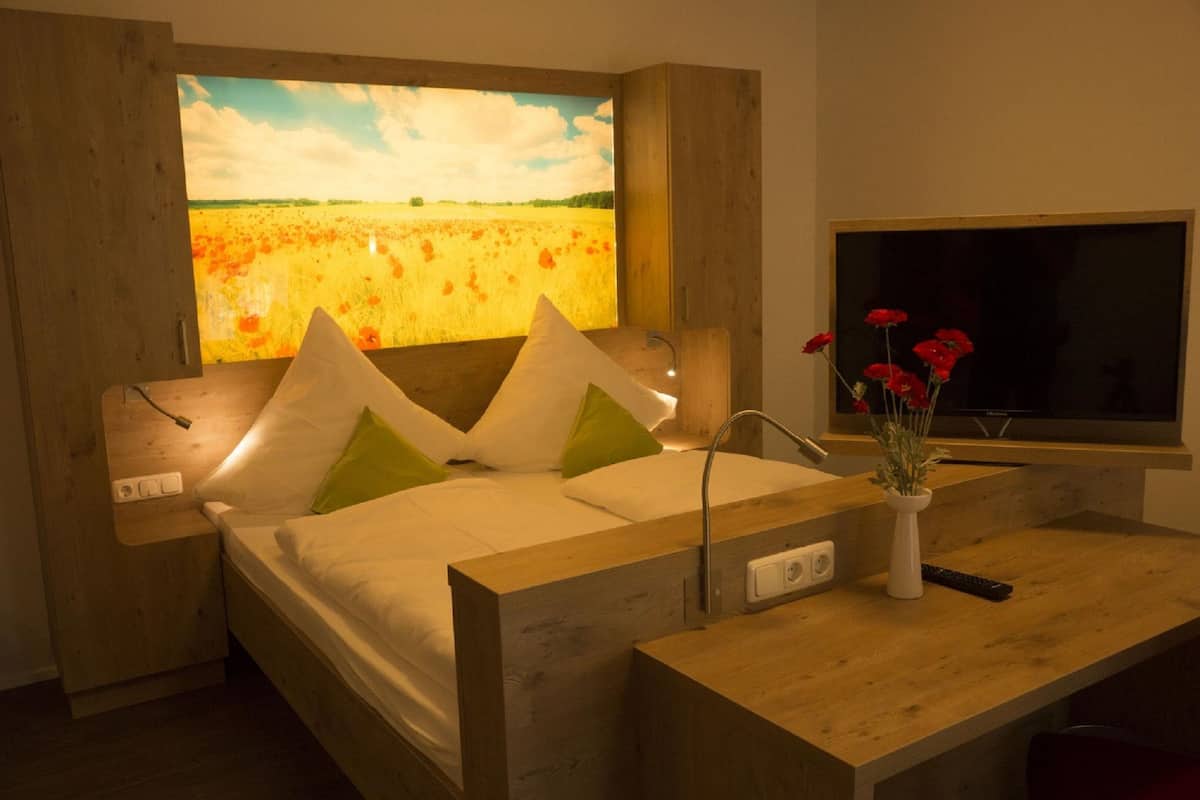 A modern bedroom with a large painting of a field, a wooden headboard, a bedside table with a vase of flowers, and a TV.