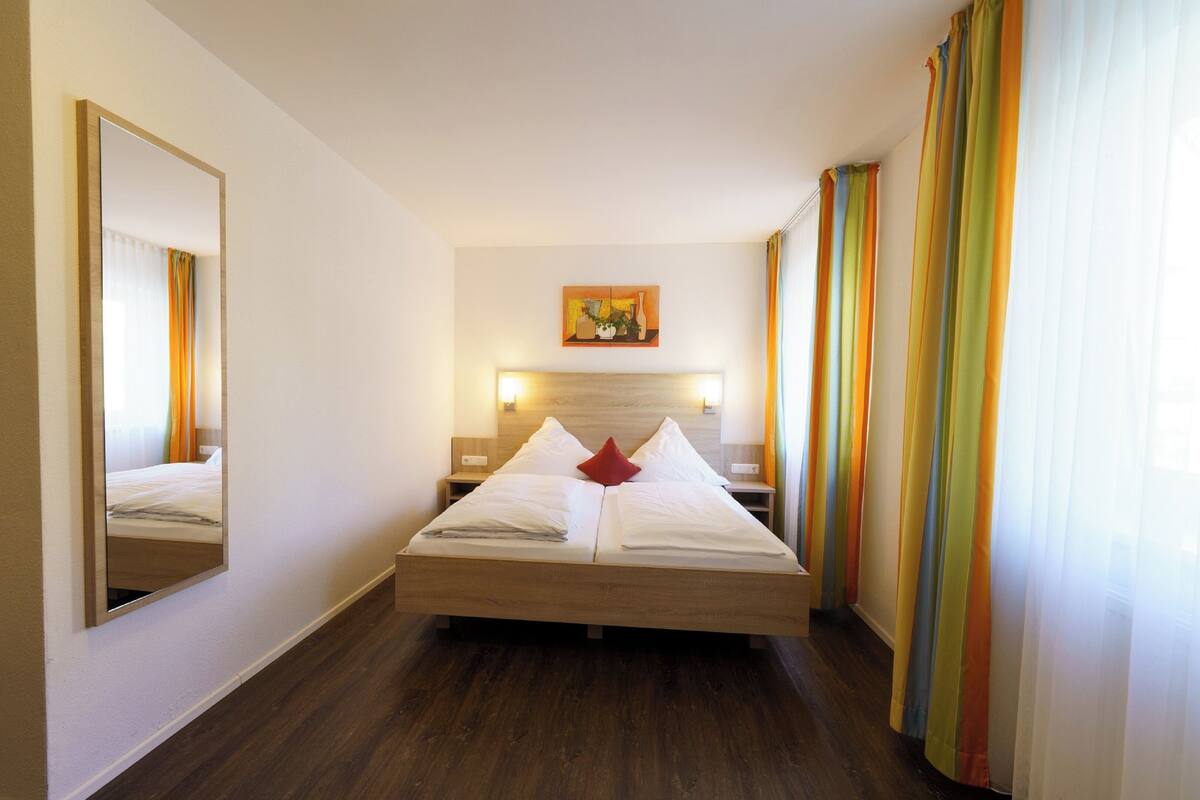 A hotel room with a large bed, a mirror, a painting on the wall, and curtains.
