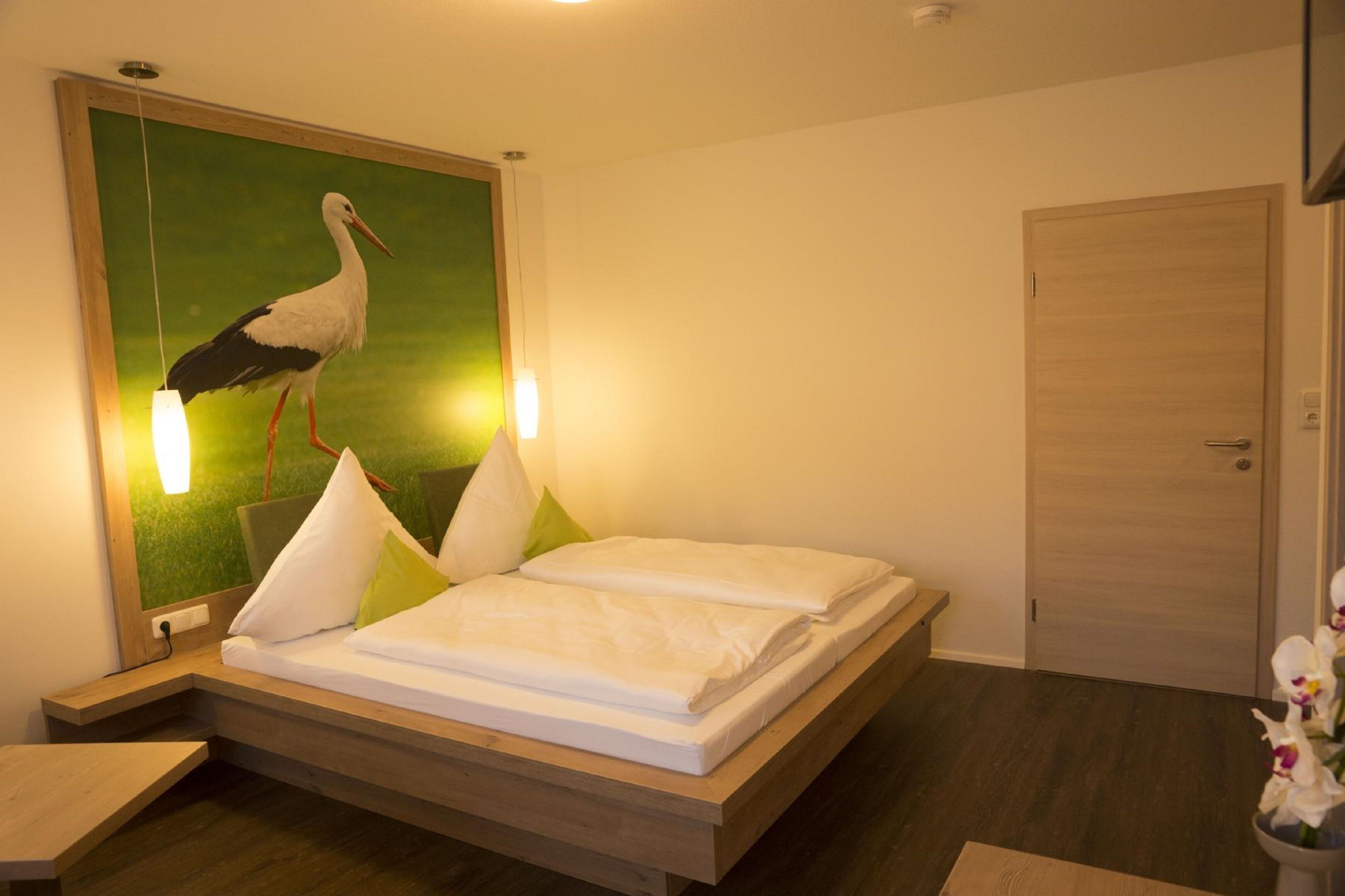 Un dormitorio con una cama, un cuadro de una cigüeña, lámparas de noche y una puerta de madera.