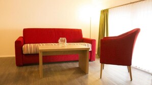 A red sofa with a striped cushion, a wooden coffee table, and a red armchair in a room with wooden flooring and a window with green curtains. - Gasthaus Schwarzer Adler (Colmberg)
