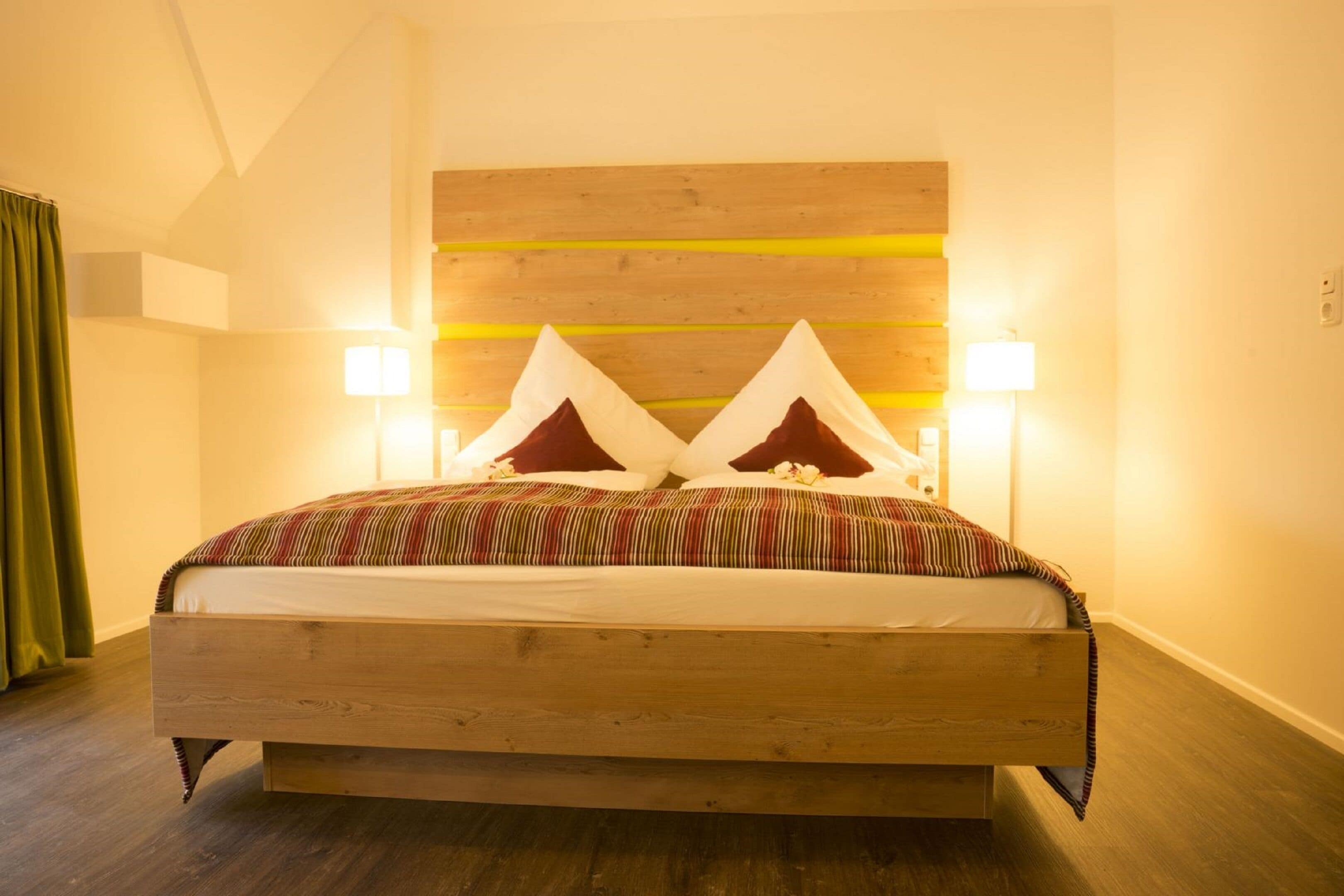 Cama com estrutura de madeira, duas almofadas e um cobertor listrado. Há duas luminárias de cabeceira e uma cabeceira de madeira com detalhe amarelo.