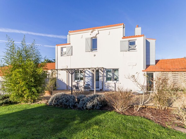 A two-story house with a covered patio area, a small garden, and a clear blue sky. - Charming Cottage near Pornic, 5km from Beach, Garden, WiFi, Family-Friendly (Pornic-Le Clion sur Mer)