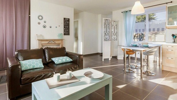 A modern living room with a brown leather sofa, a light blue coffee table, and a dining area with a white table and metal chairs. - Charming Cottage near Pornic, 5km from Beach, Garden, WiFi, Family-Friendly (Pornic-Le Clion sur Mer)