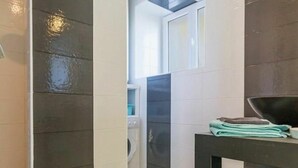 A modern bathroom with a dark tiled wall, a washing machine, and a towel rack with folded towels. - Charming Cottage near Pornic, 5km from Beach, Garden, WiFi, Family-Friendly (Pornic-Le Clion sur Mer)