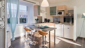 A modern kitchen with a dining area, featuring stainless steel appliances, a large window with light blue curtains, and a pendant light. - Charming Cottage near Pornic, 5km from Beach, Garden, WiFi, Family-Friendly (Pornic-Le Clion sur Mer)