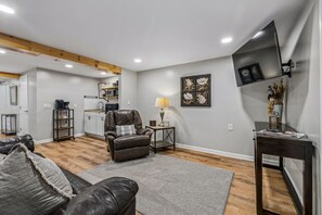 A living room with a leather sofa, a coffee table, a TV mounted on the wall, and a kitchen area in the background.