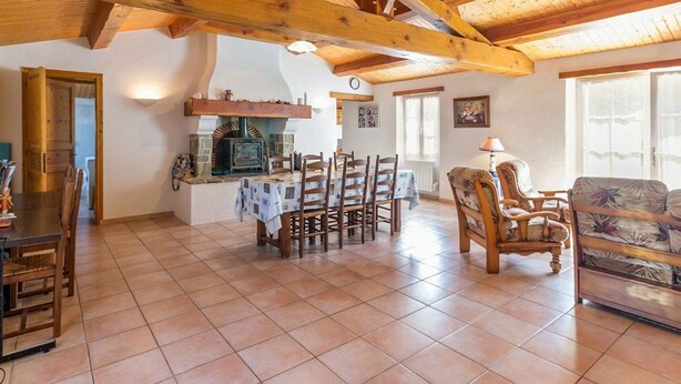 Een ruime woonkamer met een houten plafond, een stenen openhaard, een eettafel met stoelen en een bank.