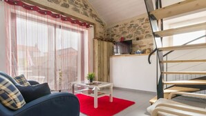 Ein gemütliches Wohnzimmer mit einem blauen Sofa, einem kleinen Couchtisch und einem roten Teppich. Eine Treppe befindet sich rechts, und hinter dem Sofa ist eine Steinwand zu sehen.