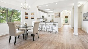 A spacious dining area with a round table and four chairs, a kitchen with white cabinets, and a view of trees through large windows.