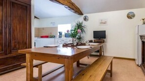 A wooden dining table with benches, a kitchen area with white cabinets, and a television on the wall. - Charming Farmhouse Near Nantes with Pond, WiFi, and Baby Equipment (Corcoué-sur-Logne)