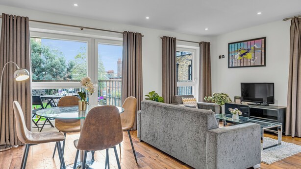 A modern living room with a grey sofa, a glass coffee table, and a TV. There is a balcony with a view of greenery and buildings. - Chic 2 Bed Apartment in Shoreditch Square (London)