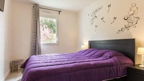 A bedroom with a purple bedspread, a dark headboard, a small bedside table with a lamp, and a window with curtains. - Modern House with Garden, Near Nantes - 3 Bedrooms, Wifi, Baby Equipment, Enclosed Garden (Nozay)