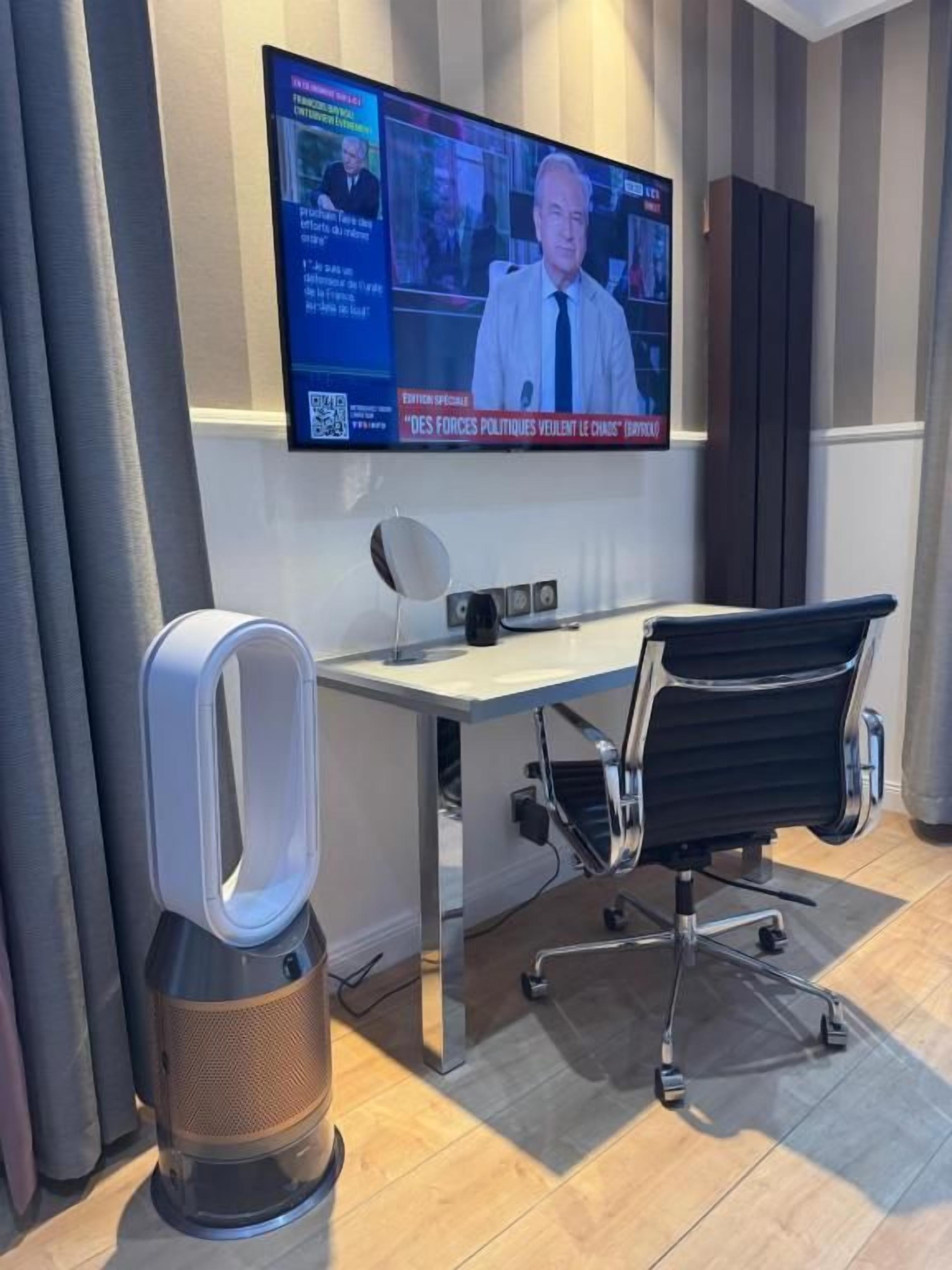 Een moderne kantoorruimte met een grote flatscreen televisie aan de muur, een bureau, een zwarte bureaustoel en een staande speaker.