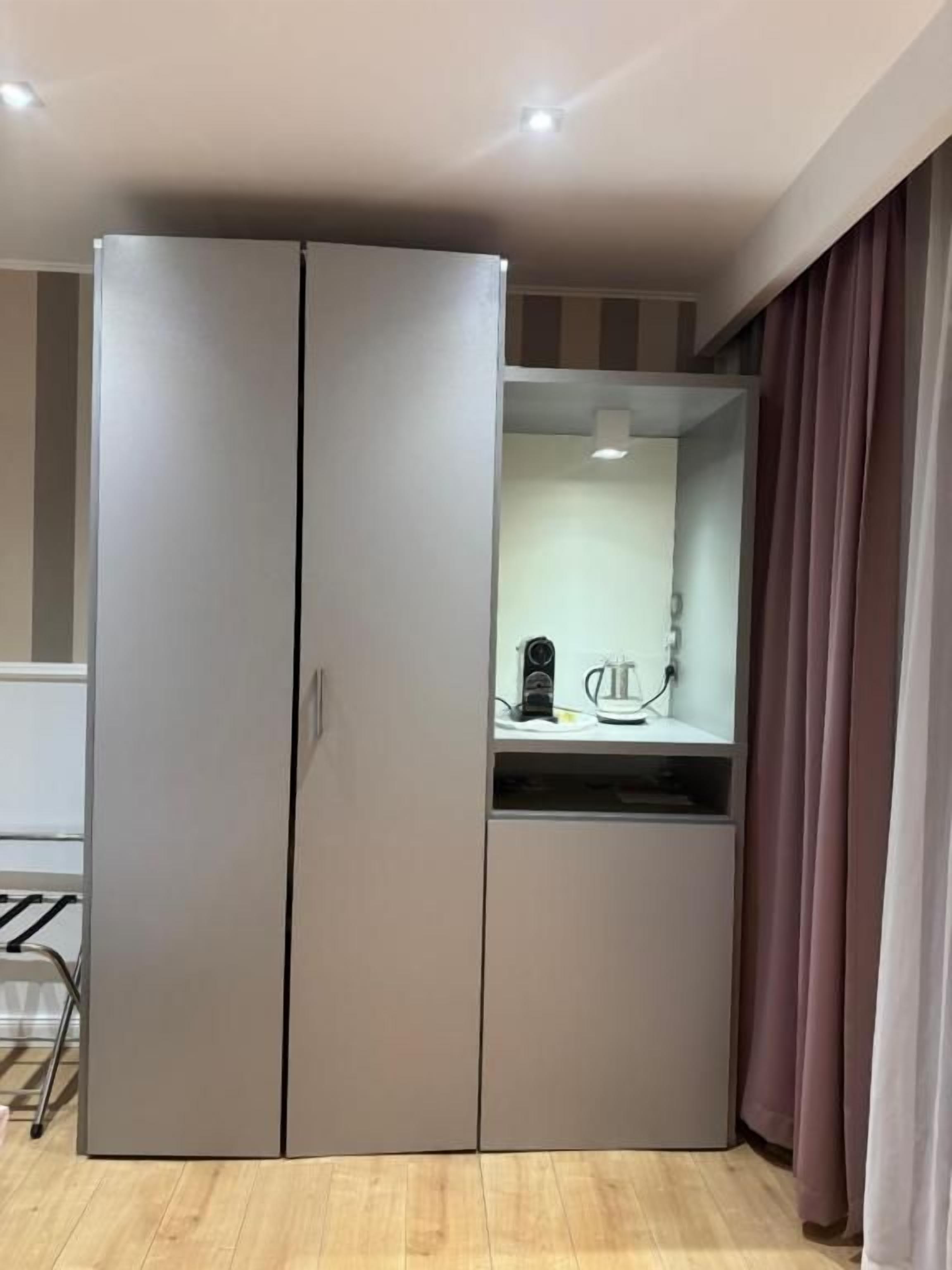 Una habitación con un armario gris, un pequeño estante con una tetera y una cortina.