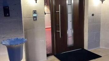 Entrada de un hotel con puerta de madera, dos apliques fijos en la pared, un teclado de seguridad y un felpudo negro.