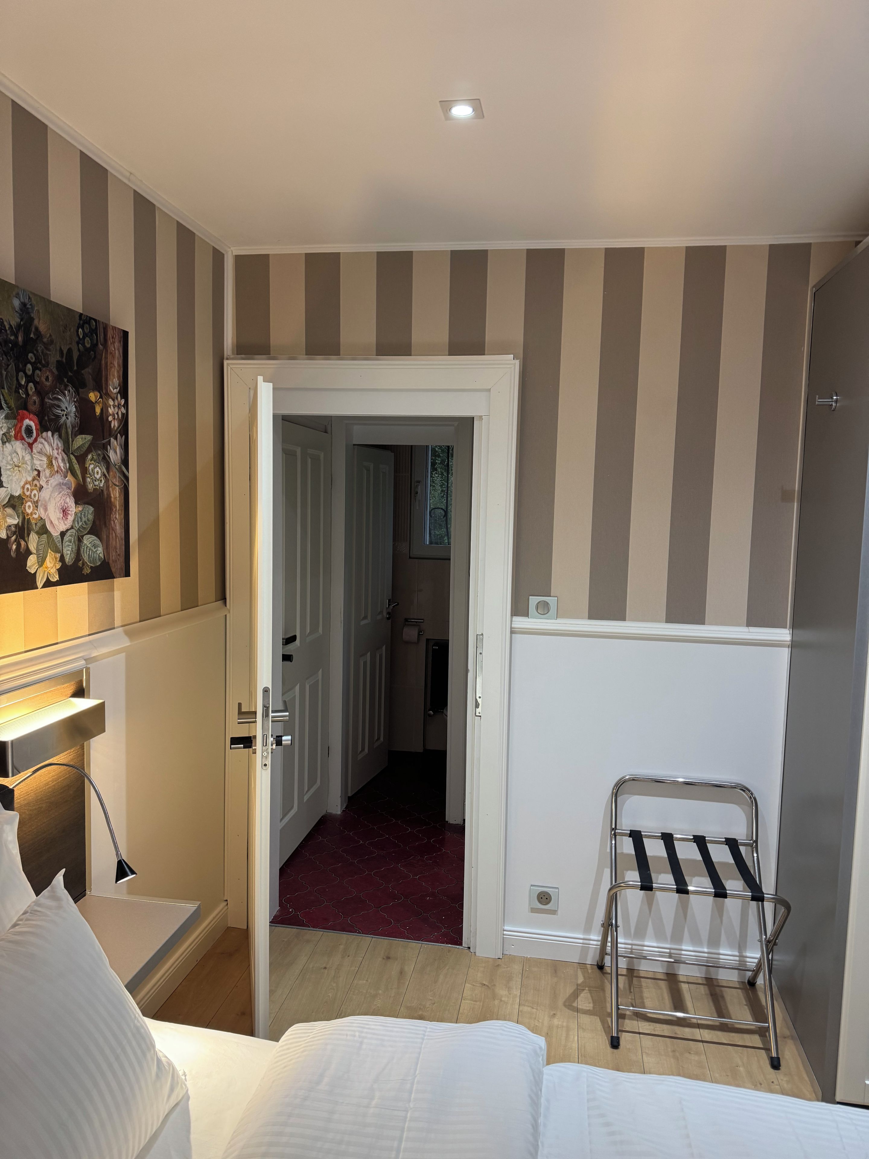 A hotel room with a bed, a desk with a lamp, a painting on the wall, and a door leading to a bathroom.