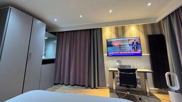 A modern hotel room with a bed, a desk with a chair, a television, and a large window with curtains.
