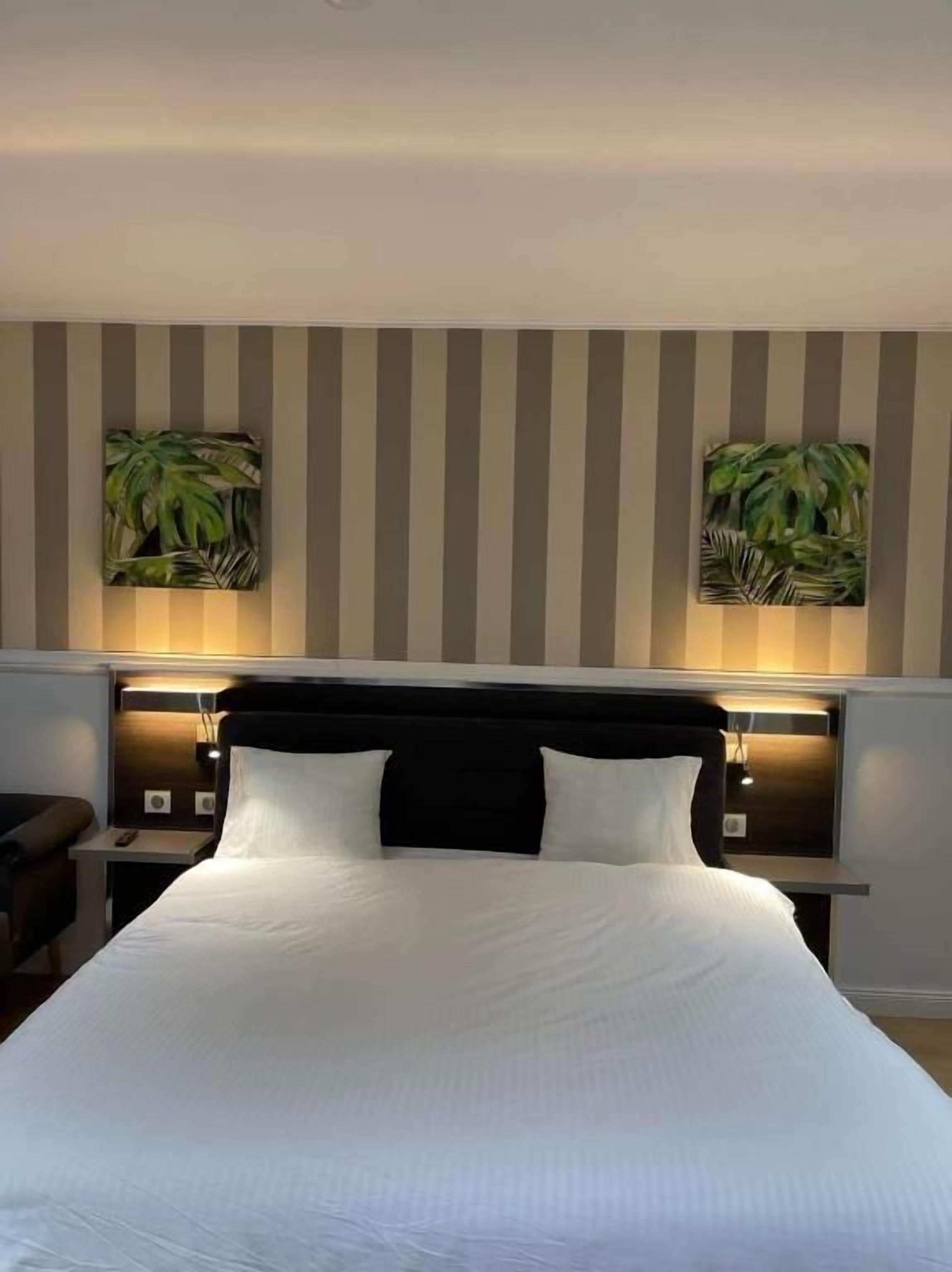 Une chambre d’hôtel avec un grand lit, deux tables de chevet, une tête de lit dotée d’un éclairage intégré et deux tableaux représentant des plantes, accrochés au mur.