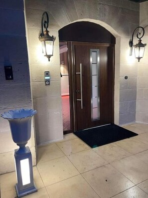 A hotel entrance with a wooden door, two wall-mounted light fixtures, a security keypad, and a black mat.