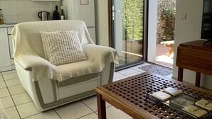Une cuisine moderne avec des meubles blancs, une chaise blanche dotée d’un coussin, une table en bois avec un plateau en verre et une porte-fenêtre coulissante donnant sur un espace extérieur.