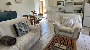 Un salon comprenant un canapé, un fauteuil, une table basse, une table à manger et un coin cuisine avec des placards et des appareils électroménagers.