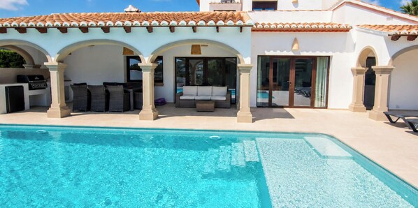A poolside area with a clear pool, outdoor seating, and a covered patio with arches.