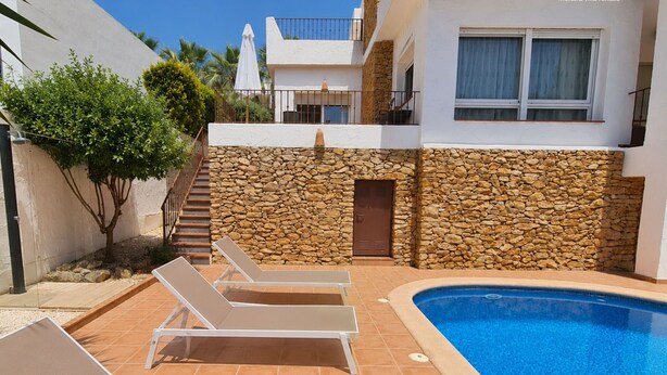 A poolside area with lounge chairs, a stone wall, and a building with a balcony.