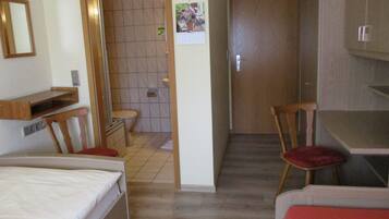 A compact hotel room with a bed, a desk, a chair, a small bathroom, and a wooden door.