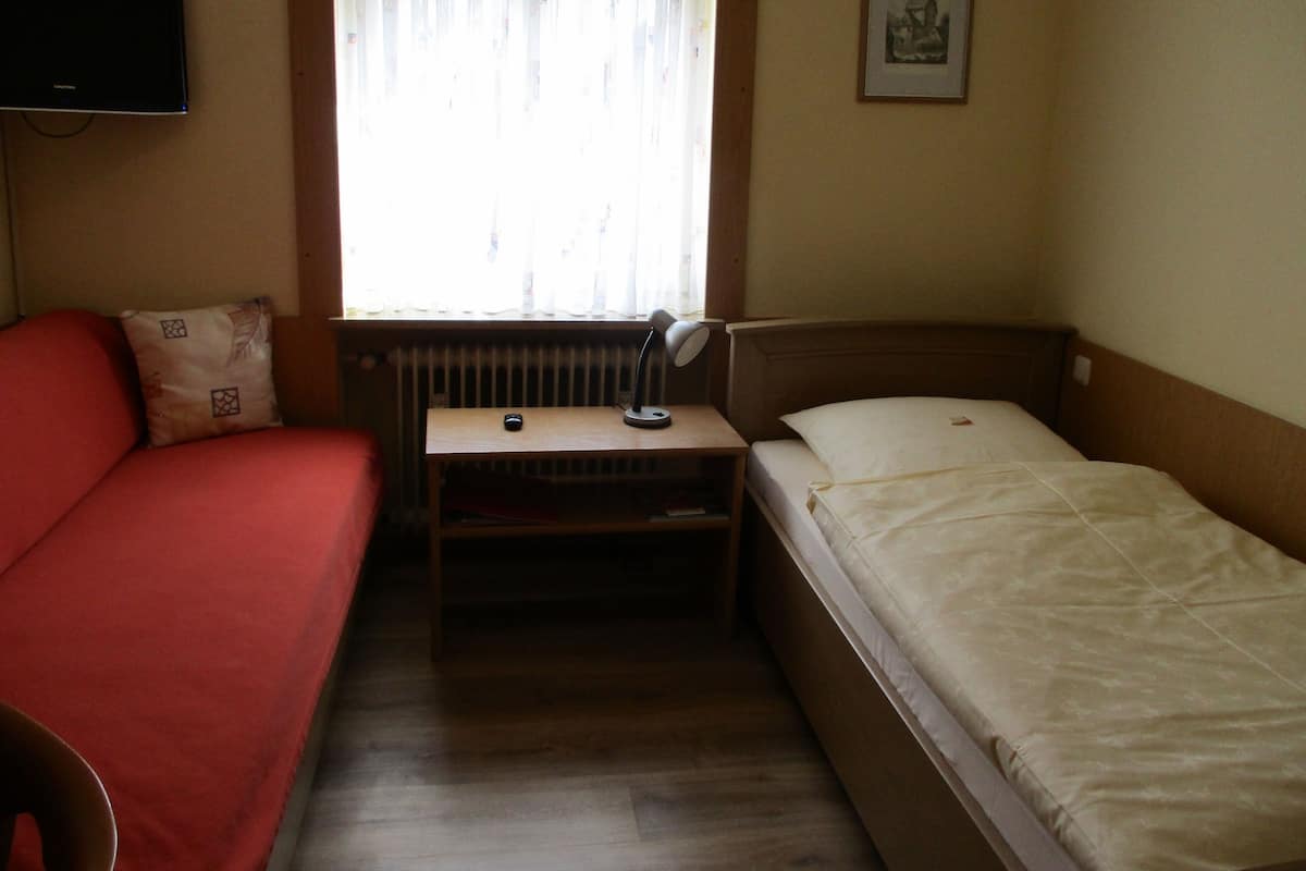 Une petite chambre d’hôtel composée d’une seule pièce, comprenant un lit, un bureau avec une lampe, une chaise, un téléviseur fixé au mur et une fenêtre avec des rideaux.