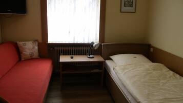 Une petite chambre d’hôtel composée d’une seule pièce, comprenant un lit, un bureau avec une lampe, une chaise, un téléviseur fixé au mur et une fenêtre avec des rideaux.