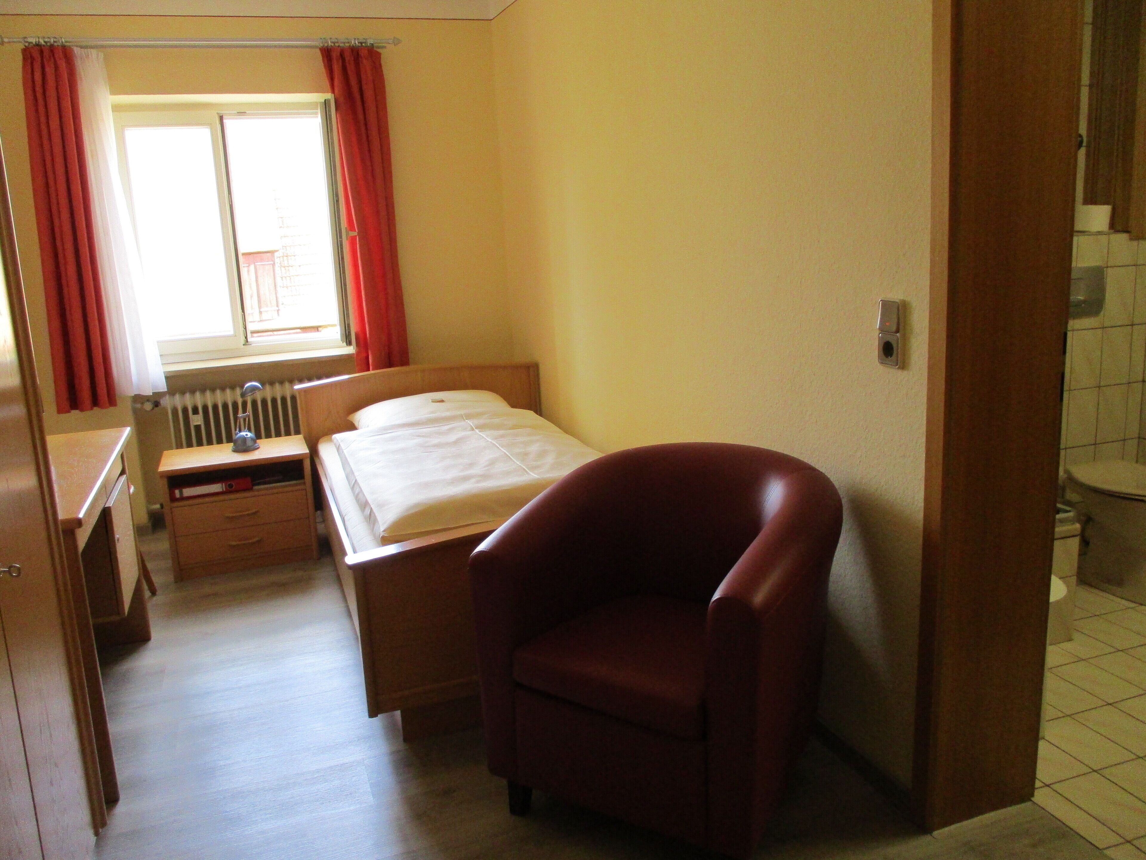 Une chambre à coucher comprenant un lit, un bureau, une chaise, un radiateur et une fenêtre avec des rideaux.