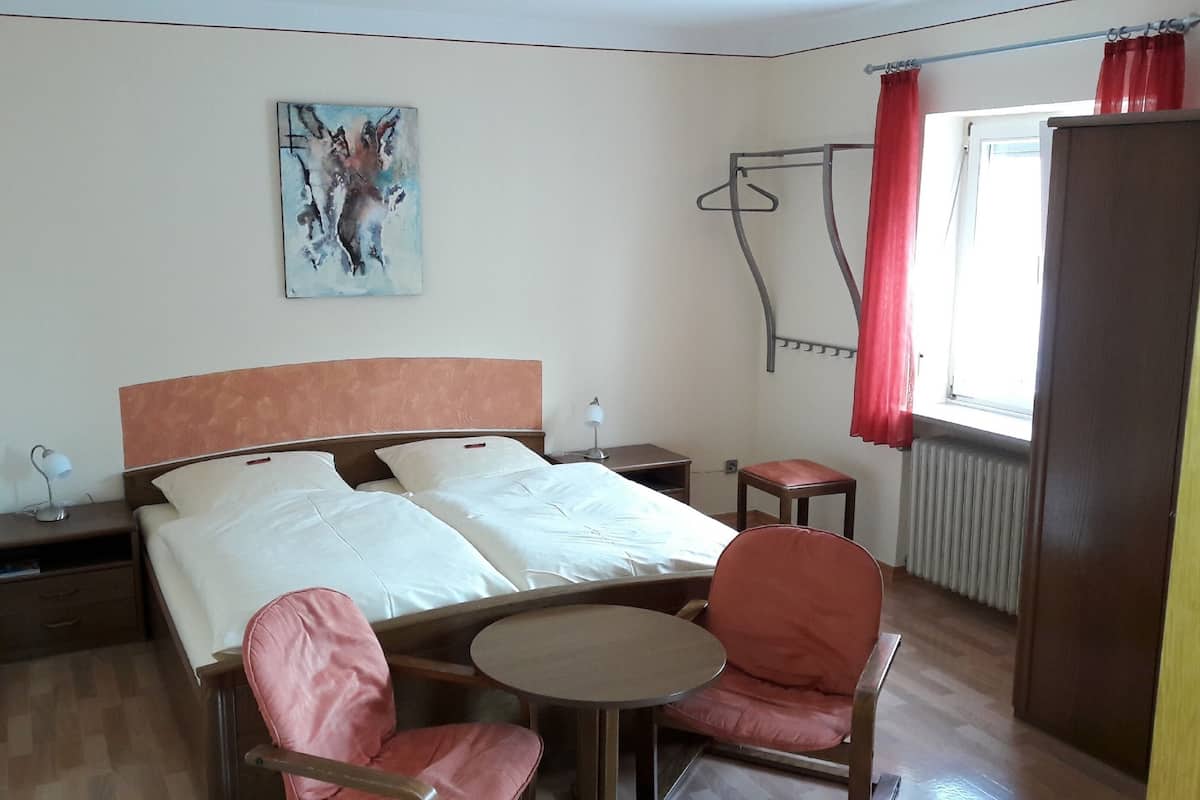 Une chambre d’hôtel comprenant un lit, des tables de chevet, une chaise, une petite table, un radiateur et une fenêtre avec des rideaux.