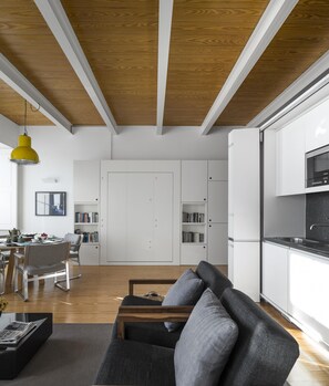 A modern kitchen with white cabinets, a black countertop, and a microwave. There is a dining area with a wooden table and chairs, and a bookshelf filled with books. - Flattered to be in Porto Sea View Apartment A (Oporto)