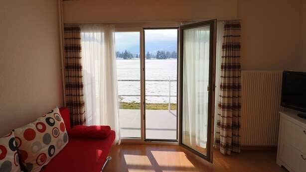 Quarto com sofá vermelho, televisão sobre um móvel branco e vista para uma paisagem nevada através de uma porta de vidro.