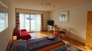 Um quarto com cama, um sofá vermelho, uma mesa de jantar com cadeiras, televisão e uma janela com cortinas.