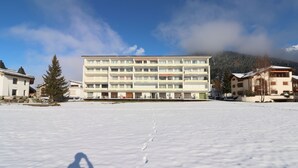 Um edifício de vários andares com telhado plano, rodeado por terreno e árvores cobertos de neve.