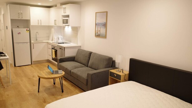 A compact hotel room with a bed, a sofa, a small table, a laptop, a refrigerator, a microwave, and a sink.