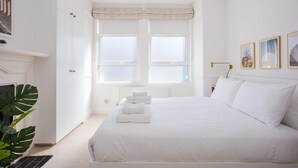 Un dormitorio blanco con una cama grande, un radiador, una ventana con cortinas, una mesita de noche y una planta.