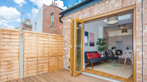 Una casa moderna con terraza de madera, puerta corrediza de vidrio que da a un espacio de estar con sofá y mesa de comedor.
