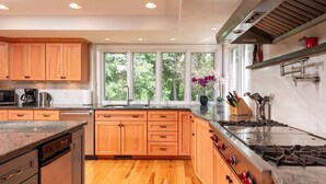 A modern kitchen with wooden cabinets, a stainless steel stove, and a large window offering a view of trees.