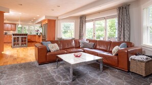 A spacious living room with a brown leather sofa, a white coffee table, and a large window with curtains.
